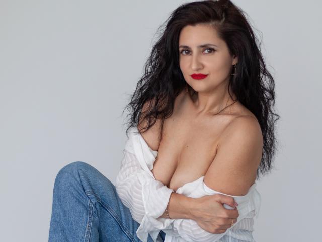 JennaEasy with wavy black hair, red lipstick, and white off-shoulder shirt, exposing her tits, sitting with arms crossed, wearing blue jeans.