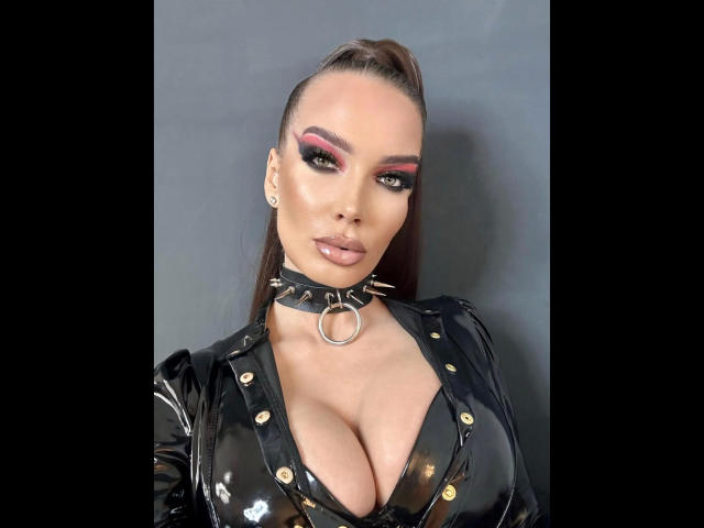 MistressKarolina-hot with big tits, wearing a black latex top, choker with a ring, pink eyeshadow, and brown ponytail, looking at the camera with a confident, sexy expression.