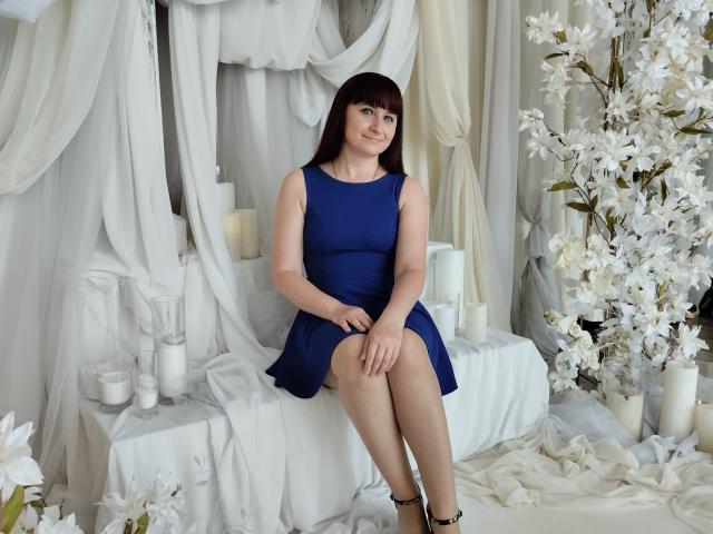 NinaFlawlees está sentada, vestida con un vestido azul, sonriendo, con las manos sobre las rodillas, rodeada de velas blancas y flores blancas. Tiene el cabello castaño oscuro y tacones negros..