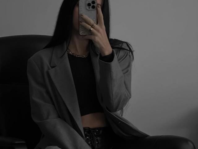 MarinaTender with long black hair, wearing a grey blazer, black top, and black leather pants, holding a phone covering her face, seated in a black chair.