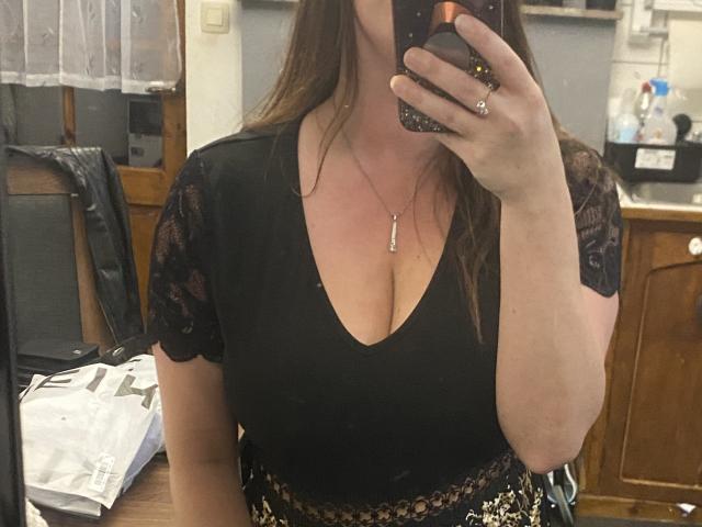 SerenaFr, con cabello largo y castaño, blusa de encaje negro, escote pronunciado que muestra un busto grande, sosteniendo un teléfono, usando una falda de estampado de leopardo, collar con un pequeño dije, anillo en el dedo..