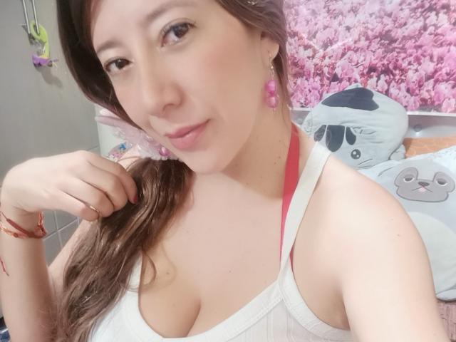 HotPamelaSex with pink earrings, brown hair, white tank top, showing her tits, pink bra strap, right hand near face, subtle smile, pink cherry earrings, bracelet, soft lighting.