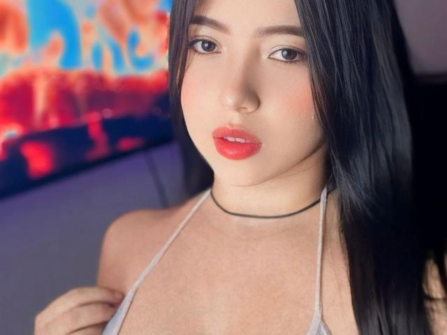 BellaDawson has long black hair, red lipstick, and a black choker. She's wearing a white halter top, showing her small tits. She looks directly at the camera.