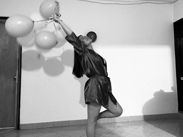 EmmaMack in a silky dress, holding balloons, one leg raised, arms stretched, shadow on wall, black and white, she's got small tits, short sleeves, tile floor.