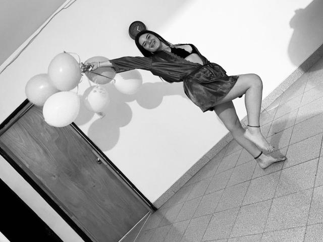 EmmaMack is leaning against a wall, holding balloons, wearing a dress, and a hat, with one leg raised, showing her bare ass and ankle bracelet in a black-and-white photo.