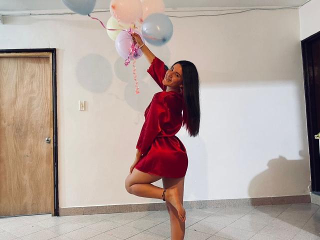EmmaMack with long dark hair, wearing a red satin robe, standing on one leg, holding pink and blue balloons, smiling, showing her bare ass, in a tiled room.