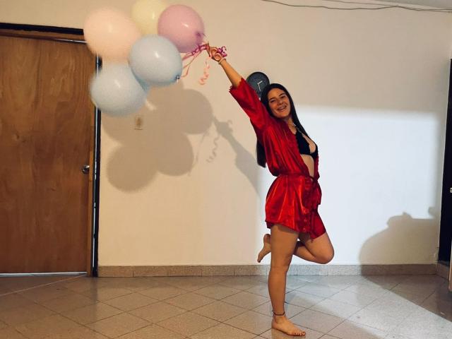 EmmaMack in a red silky robe, holding pink and white balloons, smiling, standing on one leg, with long black hair, medium tits, and a cute, happy expression.