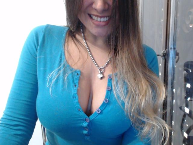 Gemma Lopez, une femme séduisante avec de longs cheveux blonds, porte un haut bleu à boutons, laissant entrevoir de larges seins, et elle sourit. Elle porte un collier avec une croix en argent, et une chaise en métal est partiellement visible sur la droite..