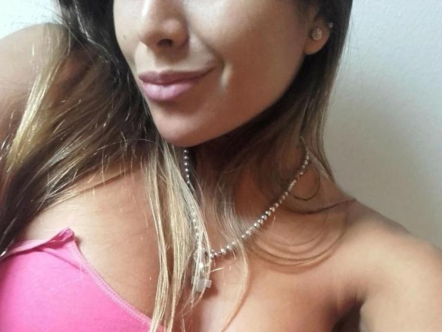 Gemma Lopez, com cabelo castanho claro, top rosa sem alças, colar de pérolas e um sorriso discreto, em um selfie, mostrando seus seios e brincos..