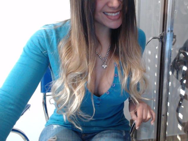 Gemma Lopez, aantrekkelijk met lang, blond ombré haar, een blauwe V-hals top, waarbij haar borsten zichtbaar zijn, lachend, met een vlinderketting om, een schaar vasthoudend, zittend op een blauwe stoel..