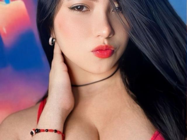 BellaDawson with long black hair, red lipstick, and a red bra, touching her neck, wearing a black choker and red bead bracelet, showing her tits.