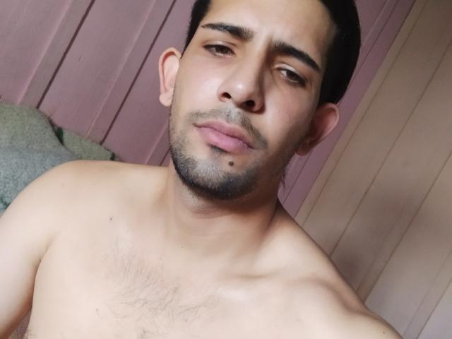 Shirtless Donino with light brown skin, dark hair, and a small mole on his chin, looking slightly bored with a subtle smirk. His chest has visible hair.