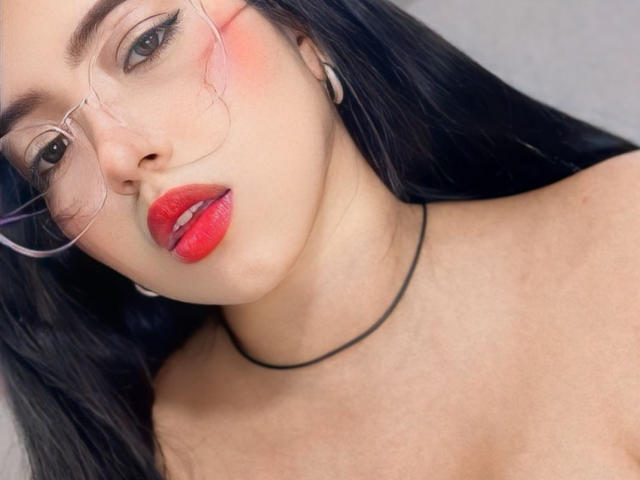BellaDawson with red lips, glasses, black hair, and a black necklace, looking at the camera with a slight pout. Her tits are not visible in the shot.
