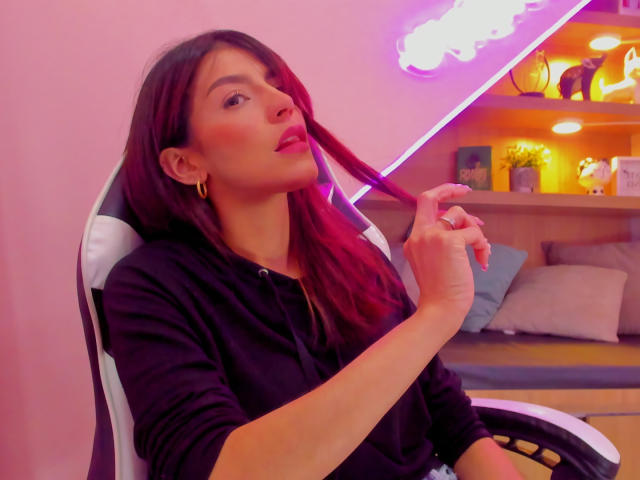 ValenRosee con cabello rojo, sudadera negra con capucha, aros dorados, sujetándose el cabello, luz neón rosa de fondo, sentada en una silla de juego blanca y negra..