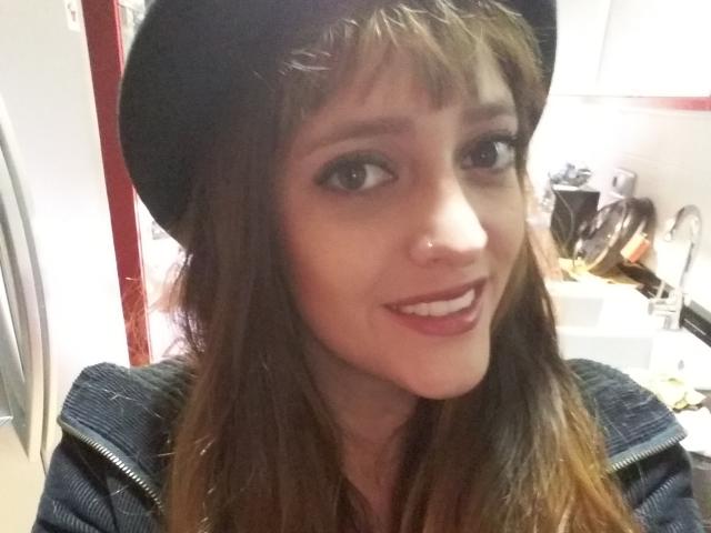 JaneRoa with brown hair, wearing a black hat, smiling, showing her teeth, nose piercing, and wearing a dark jacket, in a casual selfie. Her tits are not visible.
