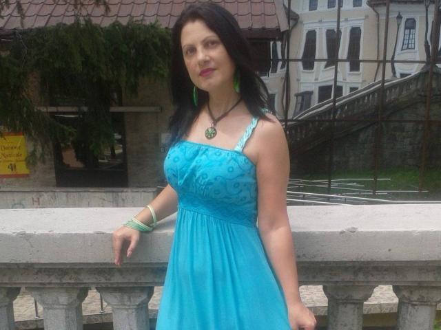 KendraSecrets with dark hair, green earrings, blue dress with floral pattern, green bracelet, necklace with green pendant, standing against white railing, looking slightly to the right.