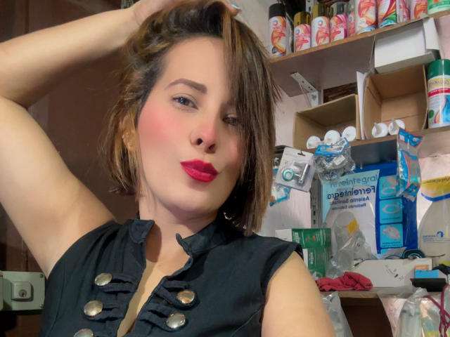 SaritaVelez with pink lips, black sleeveless top, brown hair, and raised arm, showing off her tits and a slightly blushed face, in a cluttered room.