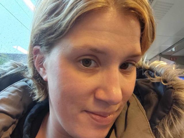 MireilleM has short blonde hair, light skin, and brown eyes. She's wearing a brown puffy jacket with a fur-lined hood. Her small nose and subtle smile are visible.