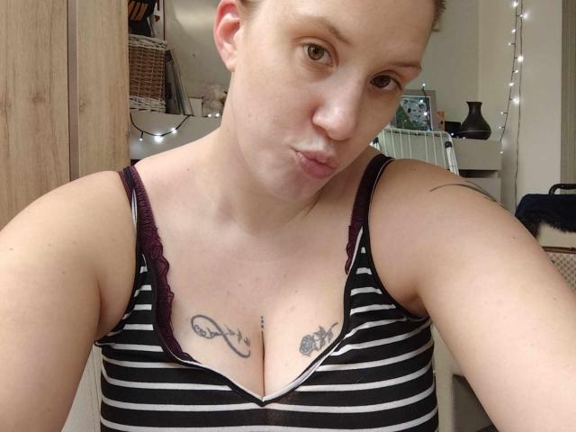 MireilleM with short brown hair, pouting lips, striped tank top, showing small tits with tattoos, black and gray ink, in a room with string lights.
