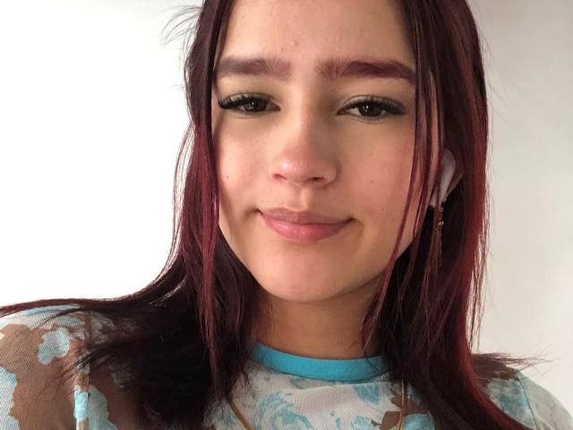 MelodyMansson has red hair, brown eyes, and a light skin tone with a subtle smile. She's wearing a blue-patterned shirt with headphones in her ears.