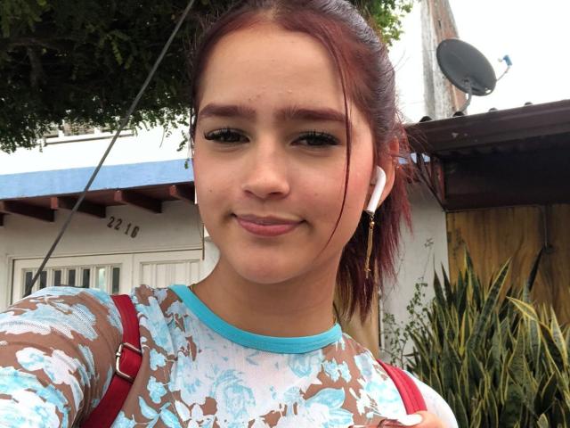MelodyMansson with red hair, blue-eyed, wearing floral shirt, earbuds, red straps, smiling, cute, small nose, light skin, medium breasts, subtle makeup, outside, daytime.