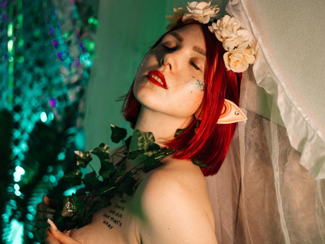 Dora, mit rotem Bob, Blumenkranz, Elfenohren, die eine Rose hält, mit grünen Blättern, rotem Lippenstift, einem Tattoo auf der Brust, mit geschlossenen Augen, weichem Licht, einem weißen Vorhang, einem grün-blauen Hintergrund..