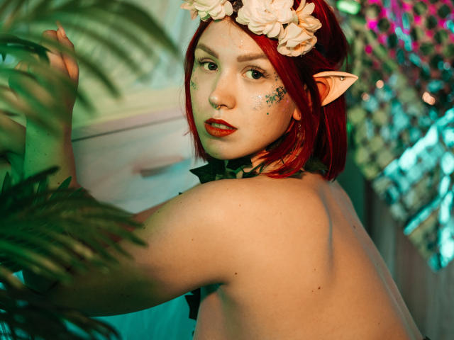 Topless DoraXDora with red hair, elf ears, flower crown, and sparkles on her face, looking over her shoulder, holding a plant, with green and pink lighting.