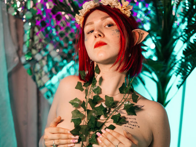Dora, mit roten Haaren, Elfenohren, einem Blumenkranz und grünen Ranken, die ihre Brüste bedecken, trägt roten Lippenstift und Stern-Tattoos und blickt mit den Händen auf der Brust nach oben..