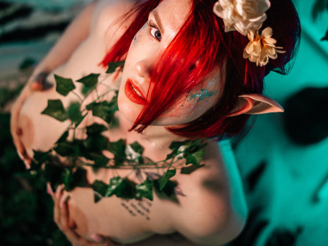 DoraXDora, oben ohne, mit roten Haaren, Elfenohren und einer Blume im Haar, hält Efeu, trägt roten Lippenstift und blauen Glitzer auf der Wange, und blickt nach oben. Brüste sichtbar..