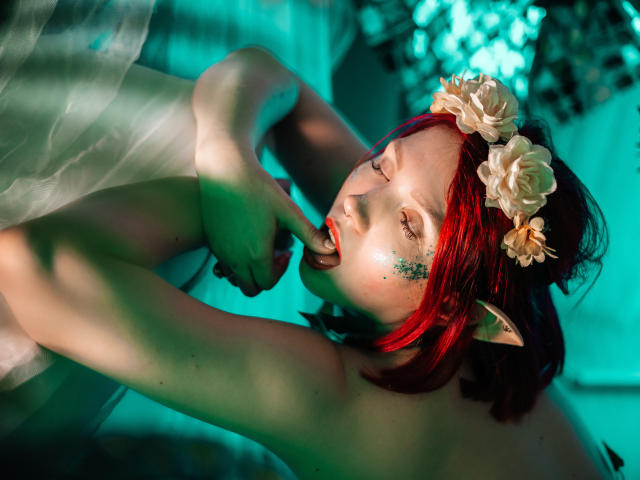 Dora, avec des cheveux roux, des oreilles d'elfe et une couronne de fleurs, touchant ses lèvres rouges avec un doigt, les yeux fermés, dans une photo de style fantastique, semi-nue, éclairée en vert..