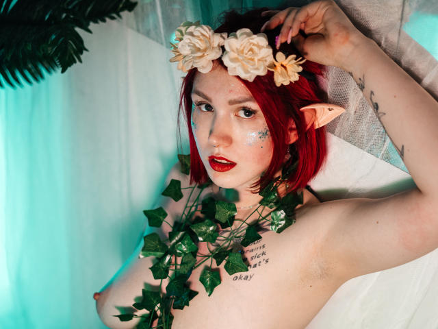 DoraXDora with red hair, elf ears, flower crown, and green vine necklace, topless, showing her tits, tattoo on right side, sparkling face, and red lipstick.