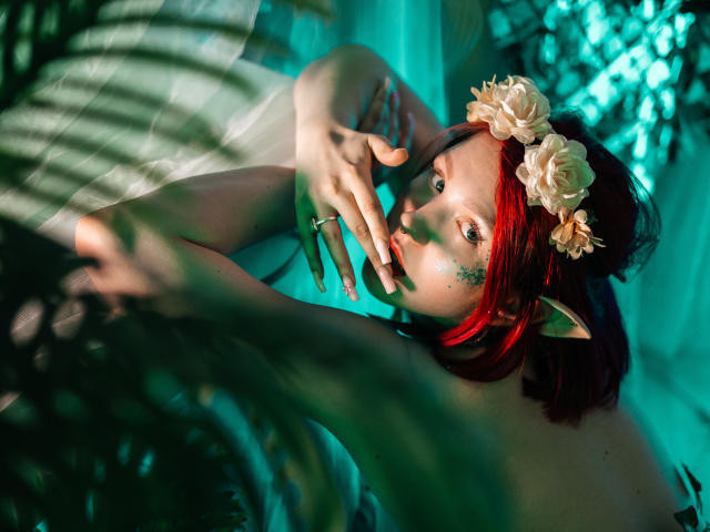 Dora, met rood haar, elfenoren, bloemenkrans, groene ogen, make-up, een ring, die haar gezicht aanraakt, met armen omhoog, teal verlichting, een wazige plant op de voorgrond, die omhoog kijkt..