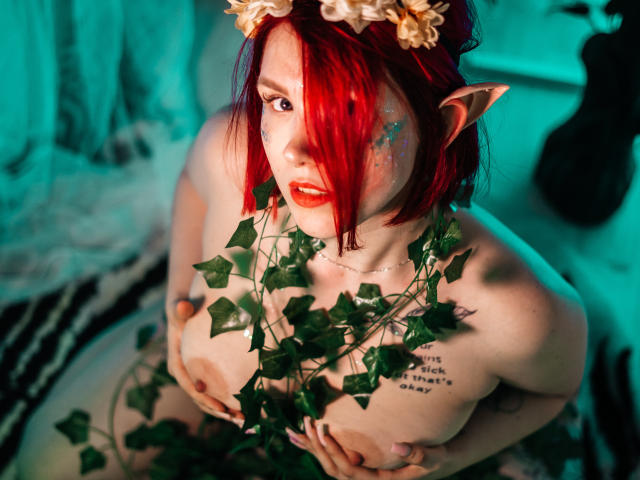 Dora, avec des cheveux roux, des oreilles d'elfe et une couronne de fleurs, tenant ses seins avec des lianes vertes, du rouge à lèvres et des tatouages, sur une photo sexy à thème fantastique, éclairée en vert..