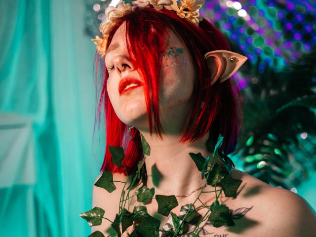 Dora: Dora mit roten Haaren, Elfenohren, Blumenstirnband, Blätterkette, rotem Lippenstift, geschlossenen Augen, Blick nach oben, strahlender Haut, sichtbarem Tattoo, grüner und blauer Beleuchtung..