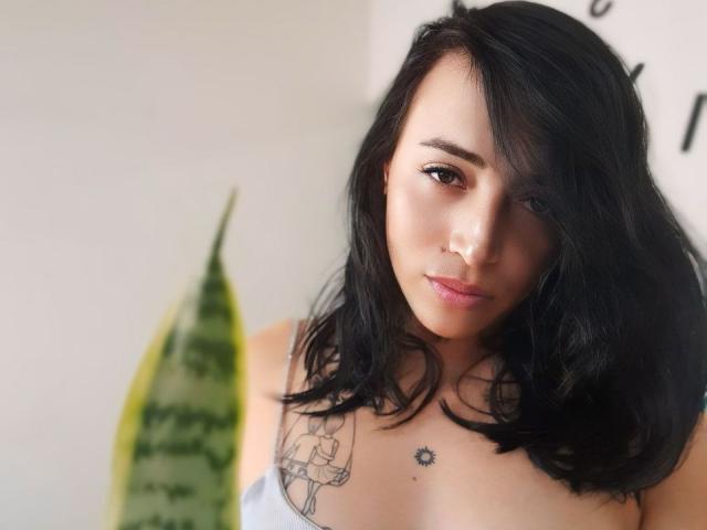YeiSela with black hair, brown eyes, and tattoos, looking at the camera, her tits visible, a green snake plant in the foreground, soft light.