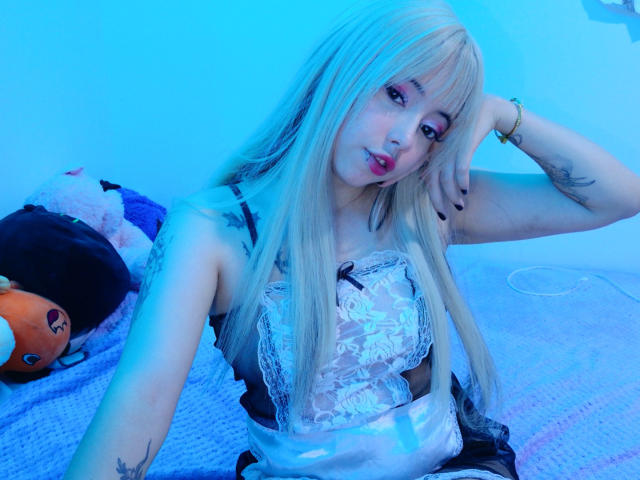Arieyaara with long blonde hair, pink lipstick, lace lingerie, and tattoos, sitting on a bed with stuffed animals, under blue lighting, looking at the camera. Her tits are visible.
