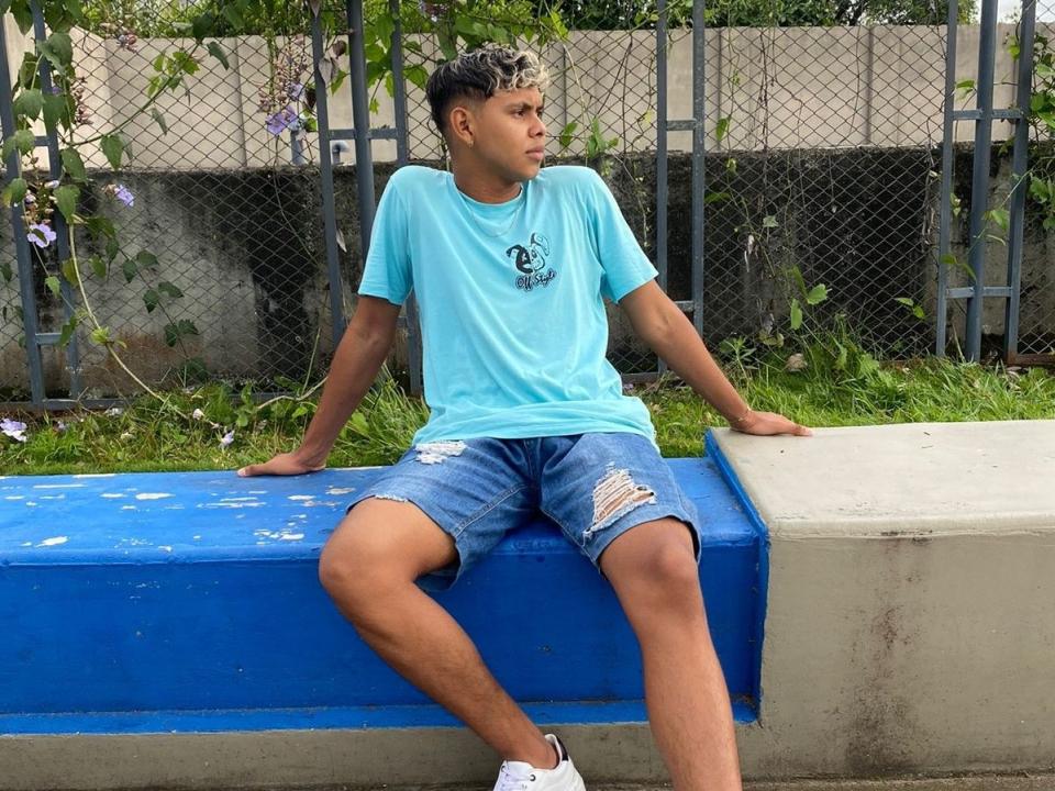 Allesso sits on a blue bench, wearing a turquoise tee, ripped denim shorts, and white sneakers, with a chain-link fence and greenery in the background.