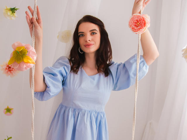 AdeleClifford with medium tits, wearing a light blue dress, holding white ropes with pink and yellow flowers, smiling, brown hair, and blue eyes, standing in a bright room.