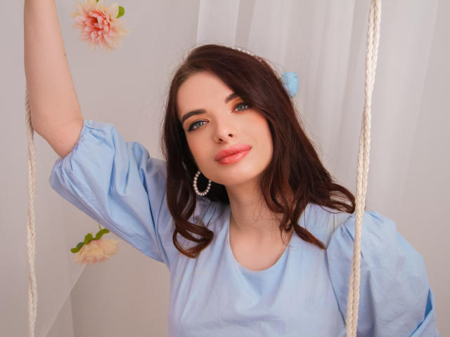 Adele Clifford, com olhos azuis, lábios rosados, cabelo castanho, vestindo uma camisa azul, brincos de argola e uma flor azul, braço levantado, cordas brancas ao fundo..