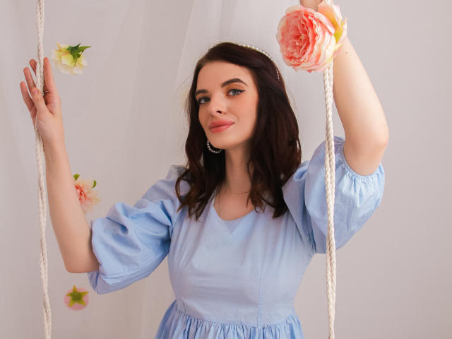 AdeleClifford with dark brown wavy hair, light blue puffy-sleeve dress, holding white ropes, pink and white flowers, smiling, fair skin, subtle makeup, pink lipstick.