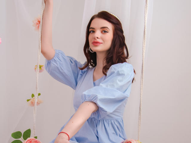 AdeleClifford with wavy brown hair, wearing a blue puffy-sleeve dress, sitting on a swing with white ropes, holding one rope, and smiling with pink lips.