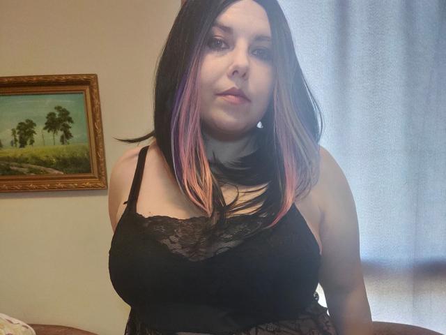 WhitteLotus has pale skin, black and pink hair, black lace top, showing her tits. She's looking at the camera with a neutral expression.