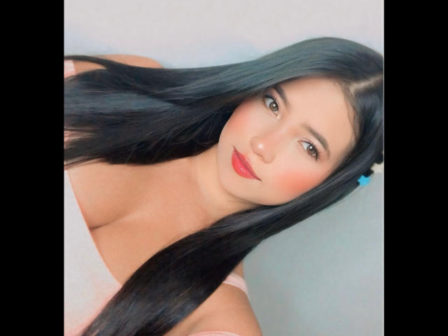 BellaDawson with long black hair, red lipstick, and pink top, showing her tits in a selfie. She has a cute face, brown eyes, and a slight smile.