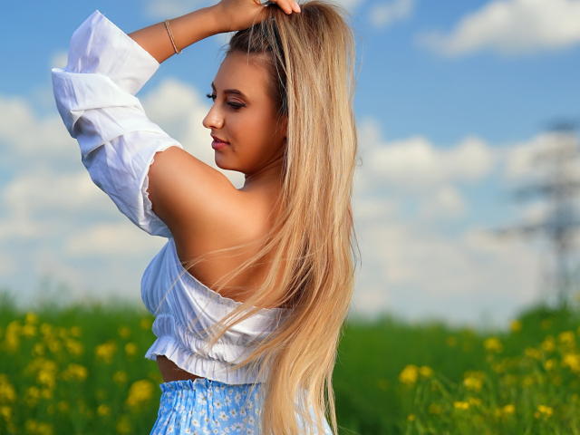 EvaRyen with long blonde hair, white off-shoulder top, and blue floral skirt, adjusting her hair, showing her bare back and ass, in a sunny field with yellow flowers.