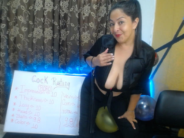 CatalinaJH with large tits in a black, open-front shirt, standing by a whiteboard, holding a pear, and smiling. She has dark hair in a ponytail, and a blue light.