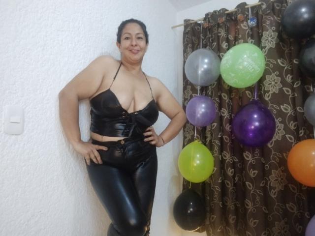 CatalinaJH with large boobs, wearing a black leather halter top and matching pants, standing against a white wall, smiling, hands on hips, with colorful balloons to the right.