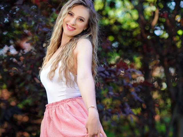 EvaRyen with long, wavy blonde hair, white tank top, pink flowy skirt, smiling, reaching out, blue eyes, fair skin, cute, standing in sunlight with bokeh background.