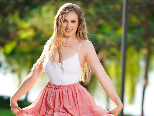 EvaRyen has wavy blonde hair, a white tank top, and a pink, flowy skirt, smiling with her hands on her skirt, showing her tits and a cute face.