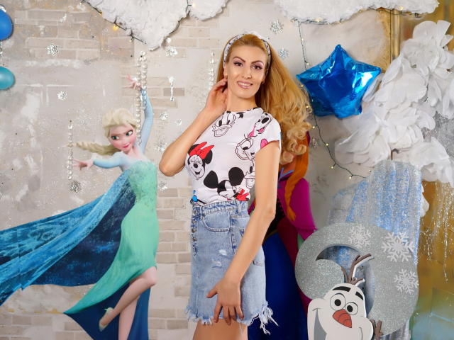 AshlineKate, con lunghi capelli biondi, indossa una maglietta con la stampa di Topolino e Minnie, una gonna di jeans sfilacciata e una fascia per capelli, sorridendo, in piedi di fronte a decorazioni a tema Frozen e innevate..