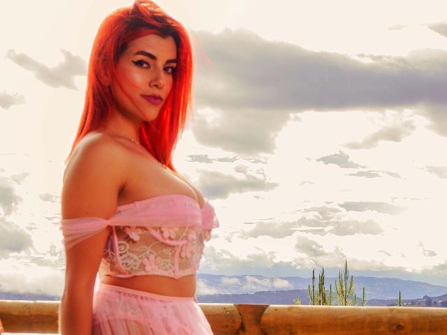 ZoeeMar with red hair, wearing a pink off-shoulder top that reveals her tits and a pink skirt, standing outside with a wooden fence in the background.